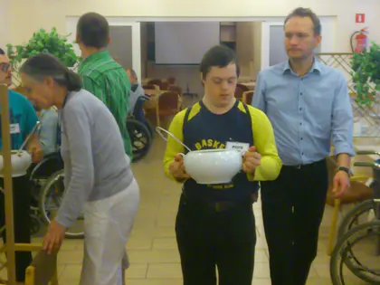 Mężczyzna z miską z posiłkiem idzie przez jadalnię, obok niego idzie opiekun