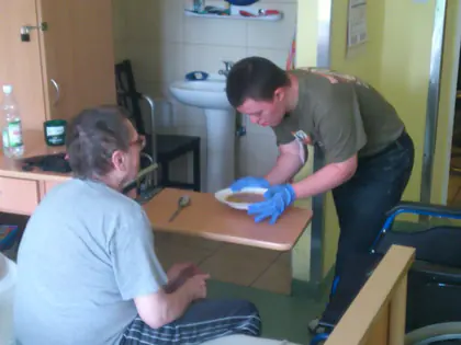 Mężczyzna w rękawiczkach podaje posiłek mieszkańcowi siedzącemu przy łóżku