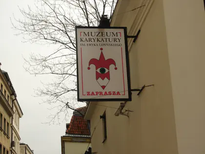 Szyld Muzeum Karykatury im. Eryka Lipińskiego z napisem Zaprasza i wizerunkiem błazna, na tle kamienicy