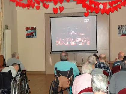 Mieszkańcy oglądają koncert wyświetlany na ekranie, nad nim wiszą czerwone papierowe serca