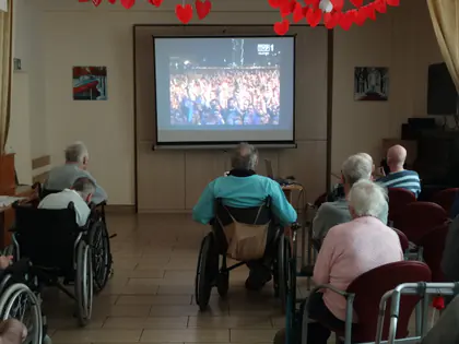 Mieszkańcy siedzący na wózkach i krzesłach oglądają na ekranie nagranie koncertu przed tłumem