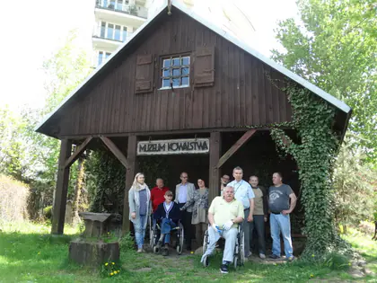 Grupa mieszkańców i opiekunów pozuje przed drewnianym budynkiem Muzeum Kowalstwa