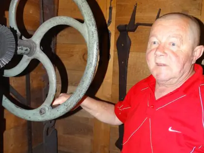 Mężczyzna w czerwonej koszulce stoi obok dużego żelaznego koła w kuźni
