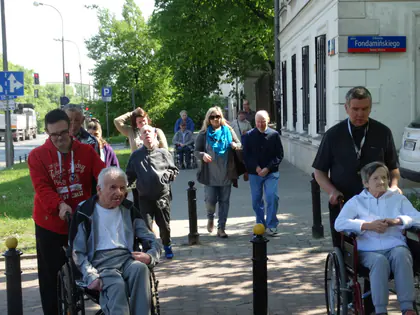 Opiekunowie pchają wózki z mieszkańcami chodnikiem przy ulicy Fondamińskiego