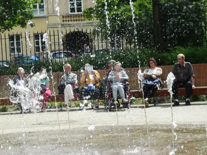 Mieszkańcy siedzą na ławkach za strumieniami fontanny w parku