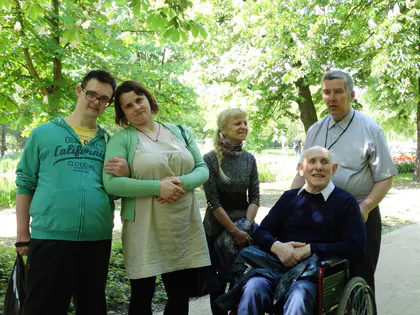 Mieszkańcy i opiekunowie pozują w parku, starszy mężczyzna siedzi na wózku