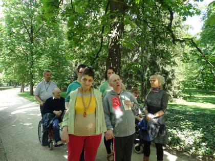 Mieszkańcy z opiekunami spacerują zacienioną alejką, mężczyzna na wózku