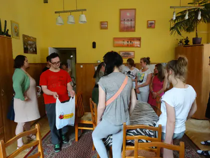 Grupa dzieci i opiekunów stoi w żółtej jadalni, chłopiec w czerwonej koszulce trzyma torbę z prezentem