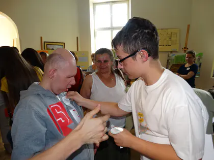 Mężczyzna w okularach przypina identyfikator mieszkańcowi w szarej bluzie, wokół stoją inni uczestnicy spotkania