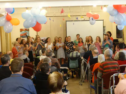 Wolontariuszki śpiewają w świetlicy udekorowanej balonami, mieszkańcy słuchają z krzeseł i wózków