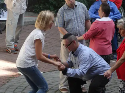 Kobieta w białej koszulce tańczy z mężczyzną w okularach, obok kibicują inni goście