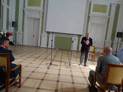 Mężczyzna w ciemnej marynarce czyta tekst przy mikrofonie w sali z kolumnami, na pierwszym planie siedzą słuchacze