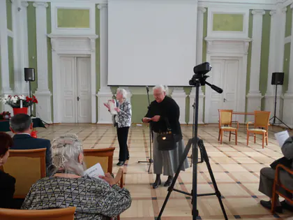 Dwie starsze kobiety stoją przy mikrofonie i kamerze na statywie, przed nimi siedzą słuchacze