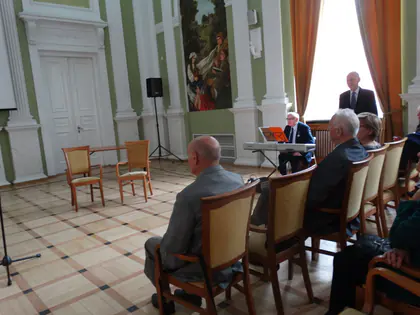 Publiczność siedzi w sali na krzesłach, w głębi muzyk przy keyboardzie i mężczyzna stojący przy oknie