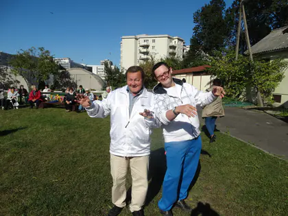 Dwóch mężczyzn w jasnych kurtkach żartuje na trawniku, w tle goście pikniku