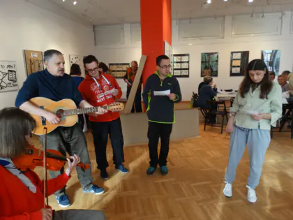 Grupa wykonawców stoi w sali wystawowej, mężczyzna gra na gitarze, obok skrzypaczka i osoby trzymające kartki z tekstem