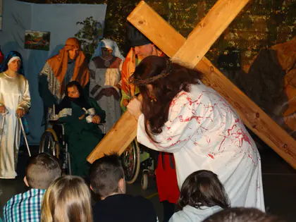 Jezus w zakrwawionej szacie upada pod krzyżem, przyglądają się postacie w kolorowych chustach