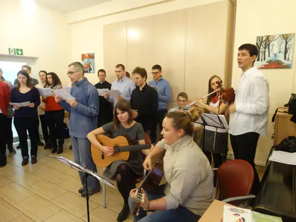 Młodzież z gitarami i skrzypcami śpiewa z kartek przed mieszkańcami w sali