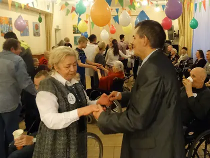 Starsza mieszkanka tańczy z mężczyzną w szarej marynarce pod kolorowymi balonami