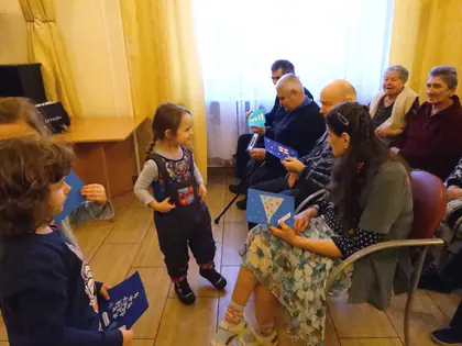 Dziewczynka podchodzi do mieszkańców siedzących pod oknem z kartkami w dłoniach