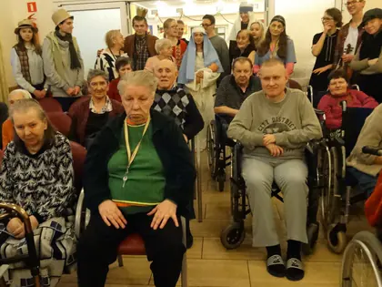 Kolędnicy w koronach i strojach pasterzy stoją za mieszkańcami siedzącymi w świetlicy