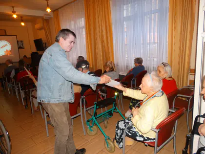 Mężczyzna wręcza kartkę starszej kobiecie siedzącej z balkonikiem w sali