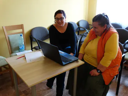 Prowadząca i mieszkanka w pomarańczowej kamizelce uśmiechają się przy laptopie
