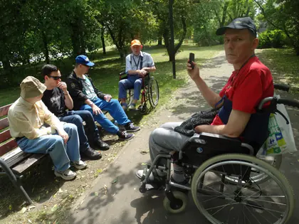 Mieszkańcy odpoczywają na ławkach w parku, mężczyzna na wózku robi zdjęcie telefonem