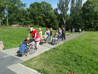Opiekunowie pchają wózki z mieszkańcami alejką przez zielony park