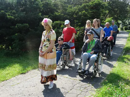 Młoda kobieta w kwiecistej sukience i chuście idzie przed grupą osób na wózkach