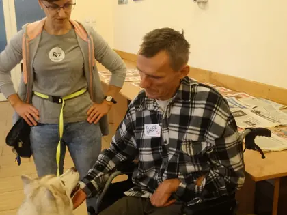 Mieszkaniec na wózku głaszcze psa husky, obok stoi prowadząca ze smyczą