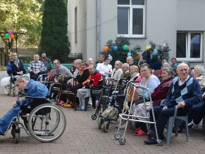 Mieszkańcy na wózkach i krzesłach siedzą w rzędach na podwórku podczas festynu
