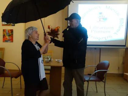 Mieszkanka z czarnym parasolem i mieszkaniec w czapce odgrywają scenkę przed ekranem
