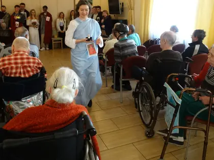 Młoda kobieta w stroju świętej idzie między mieszkańcami siedzącymi w świetlicy
