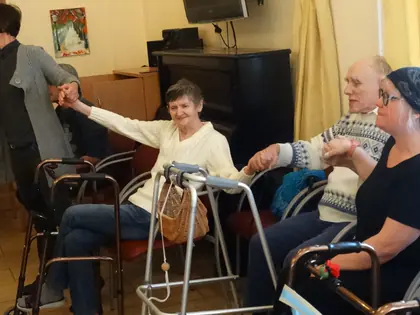 Kobieta trzyma za ręce mieszkańców siedzących przy balkonikach, wszyscy kołyszą się w tańcu