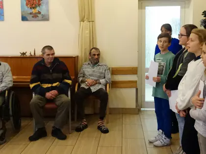 Trzech mieszkańców siedzi pod ścianą, obok stoją dzieci z kartkami