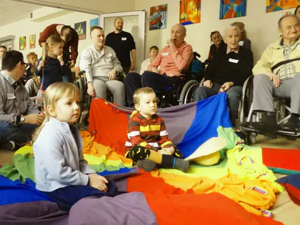 Małe dzieci siedzą na kolorowej chuście animacyjnej przed mieszkańcami
