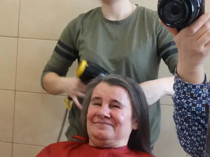 Uśmiechnięta mieszkanka w czerwonej pelerynie, za nią fryzjerka z suszarką