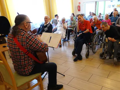Gitarzysta w kraciastej koszuli gra przed mieszkańcami na wózkach