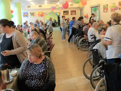Mieszkańcy siedzą przy stołach podczas jarmarku, opiekunki rozlewają napoje