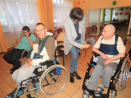 Kobieta z mikrofonem rozmawia z mieszkańcami na wózkach w świetlicy