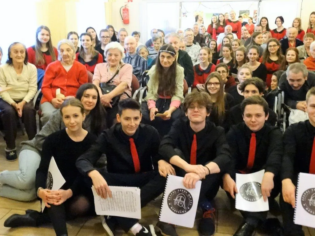 Mieszkańcy i młodzież w czerwono-czarnych strojach na wspólnym koncercie w świetlicy.