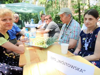 Mieszkańcy przy stole z tabliczką DPS Wójtowska na pikniku