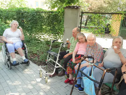 Mieszkańcy z balkonikami siedzą przy ogrodzeniu w cieniu