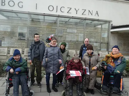 Mieszkańcy na wózkach z opiekunami przed ścianą z tablicami pamiątkowymi przy sanktuarium