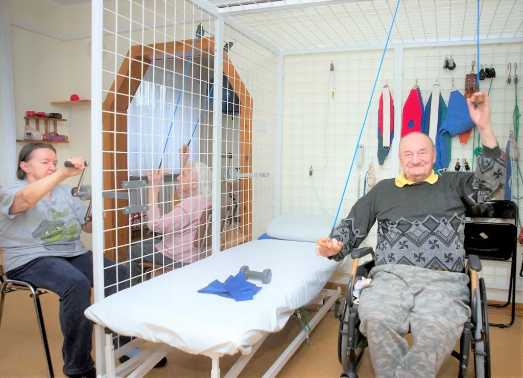 Mieszkańcy ćwiczą w sali rehabilitacyjnej z użyciem wyciągów i ciężarków.
