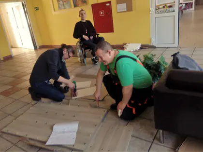 Mężczyźni składają meble na podłodze holu, obok mieszkaniec na wózku