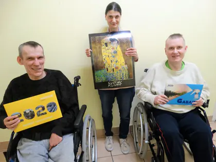 Dwaj mieszkańcy na wózkach i mieszkanka prezentują podarowane kalendarze na 2020 rok