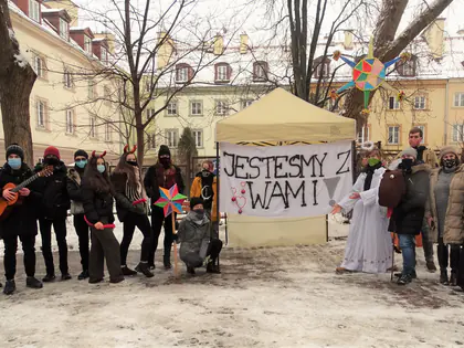 Uczniowie w przebraniach kolędników z gwiazdą i gitarą stoją przy transparencie Jesteśmy z Wami