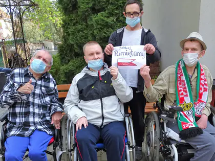Mieszkańcy w maseczkach, część na wózkach, trzymają kartkę z napisem 3 maja Pamiętamy